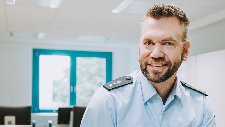 Das Headerbild zeigt einen Bundespolizisten in Uniform, der freundlich in die Kamera lächelt. Im Hintergrund sieht man einen Konferenzraum mit Fenstern, die natürliches Licht einlassen. Der Artikel beschäftigt sich mit dem mündlichen Testverfahren der Bundespolizei.